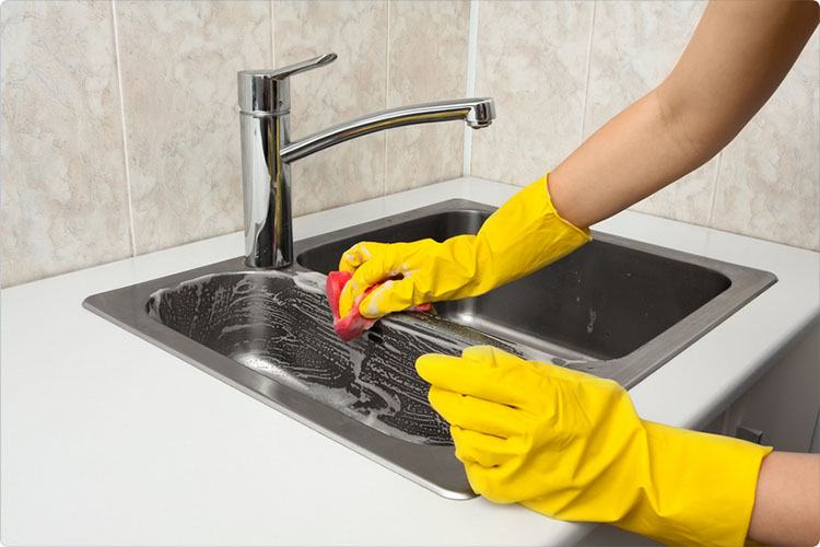 A woman shows how to clean a kitchen sink with gloves and a sponge